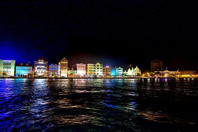 Illuminated buildings by river against sky at night