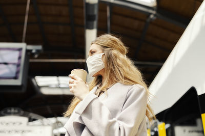 Low angle view of woman wearing mask holding coffee while standing at railroad station