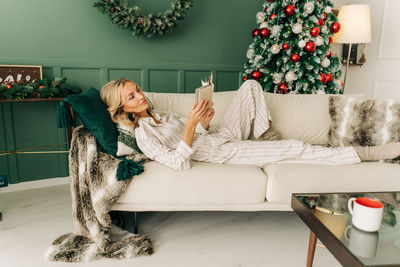 A blonde woman is lying on a cozy sofa in a living room decorated for christmas reading a book.