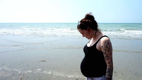 Side view of pregnant young woman standing at beach against clear sky during sunset