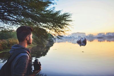 Man holding camera while standing by lake against sky during sunset