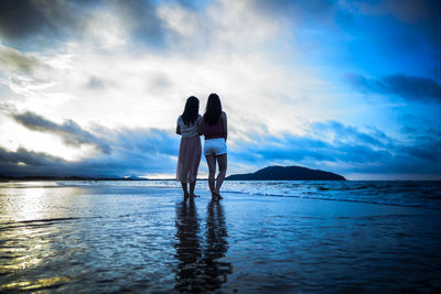 Rear view of friends standing at beach against cloudy sky during sunset