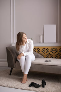 A young woman is sitting on the sofa with glasses upset, covering her face with her hand. 