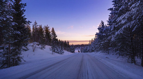 Road amidst snow covered trees against sky during sunset