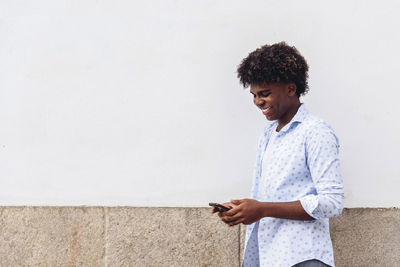 Side view of smiling african american male standing near urban building in city and surfing internet on cellphone