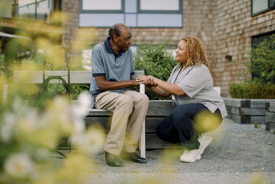 Full length of female nurse squatting while talking to retired senior man sitting on bench at back yard