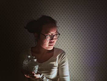 Woman wearing eyeglasses holding jar against wall at home