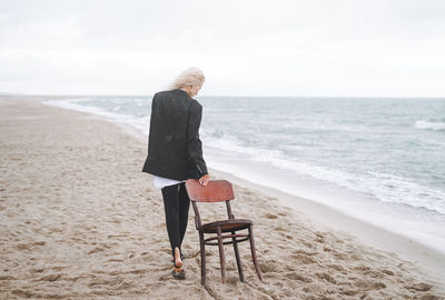 Portrait of elegant blonde woman in black suit with vintage wooden chair by sea in a storm