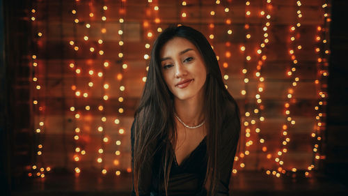 Portrait of young woman standing against illuminated string lights at night