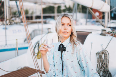 Beautiful blonde young woman in blue dress with glass of soda on boat at pier in sunset time