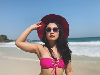 Portrait of woman in bikini wearing hat and sunglasses while standing at beach