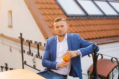 Young handsome man is sitting in an outdoor summer cafe in a blue suit and holding a cocktail glass