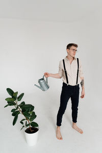 Young man looking away while watering plant against white background