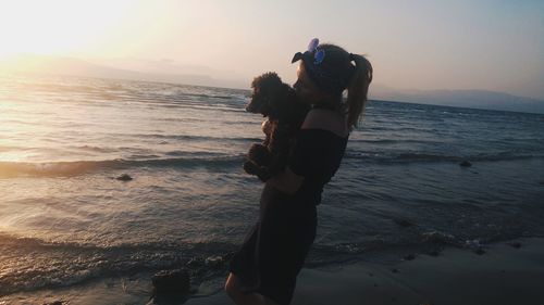 Woman with dog standing at beach against sky during sunset