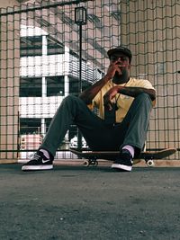 Full length of young man sitting on skateboard against metallic fence