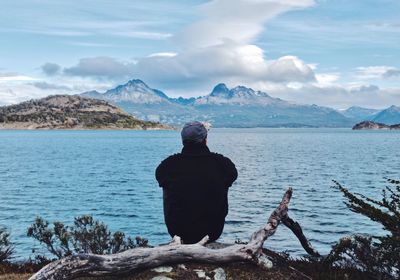 Rear view of man sitting at lakeshore against cloudy sky