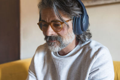 Mature man with a beard, glasses and headphones, sits on the sofa at home and works with a tablet 