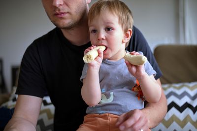 Little toddler eating banana held by his father