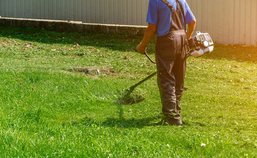 Worker man in protective pants mows green grass on lawn with grass trimmer.