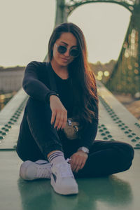 Portrait of young woman wearing sunglasses while sitting on the bridge