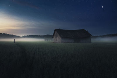 Person standing on field by barn against sky during foggy weather at night