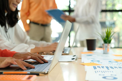 Midsection of women using laptops on desk while male colleagues discussing in background