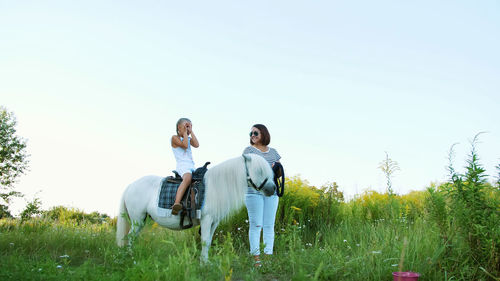 Mom and daughter are walking around the field, daughter is riding a pony, mother is holding a pony