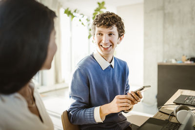 Happy young entrepreneur holding smart phone discussing with colleague in creative office