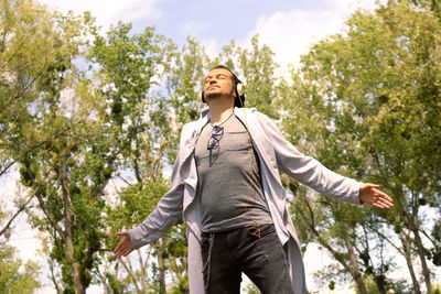 Man listening music over headphones while standing with arms outstretched at the park.