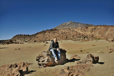 Full length of man sitting on rock at volcanic landscape against clear blue sky during sunny day