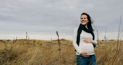 Smiling pregnant young woman touching abdomen while standing on grass against sky