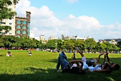 Friends lying on field at park against sky in city
