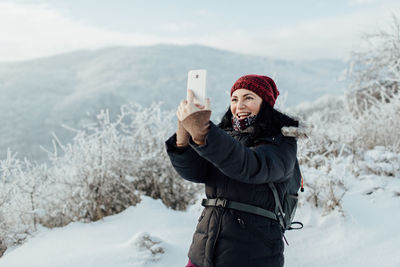 Smiling woman in warm taking selfie while standing on snowcapped mountain