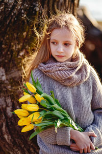 Cute girl holding flowers standing by tree outdoors