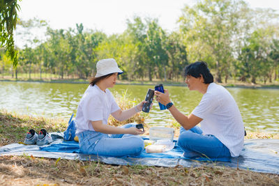 Couple toasting water bottles while sitting against lake