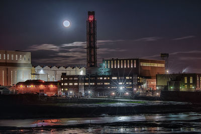 Illuminated factory by city against sky at night