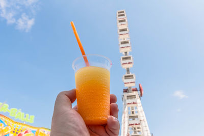 Cropped hand holding juice glass against ferris wheel in city