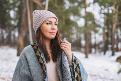 Portrait of young brunette beautiful woman in hat and grey poncho in the winter forest