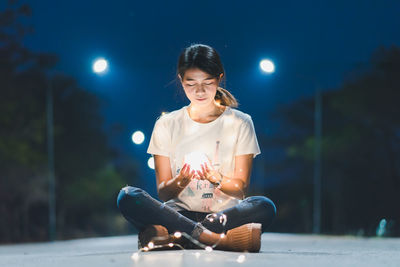 Young woman holding illuminated lighting equipment while sitting on road against trees at night