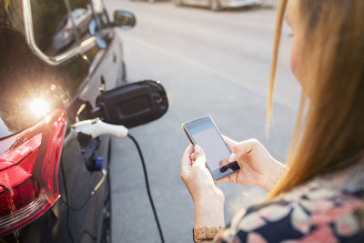 Woman using mobile phone while standing by charging electric car