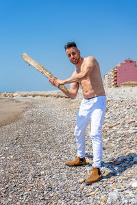 Full length of shirtless man holding wood while standing at beach