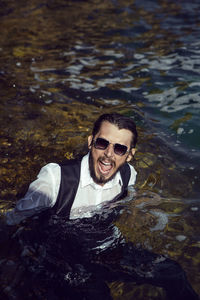 Man with a beard and sunglasses in clothes a vest and a white shirt swims in the sea among the rocks