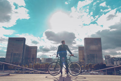 Rear view of man with bicycle standing on bridge in city against cloudy sky