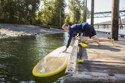 Side view of man touching paddleboard while leaning on pier