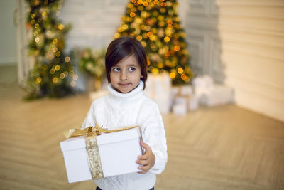 Portrait of a boy a child in a white sweater holding a gift box at the christmas tree