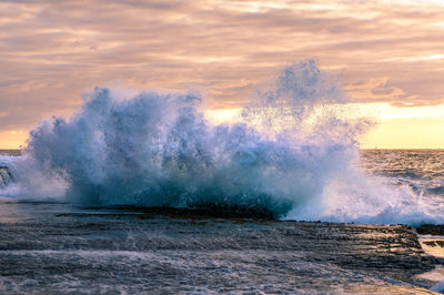 Sea waves splashing on shore against sky during sunset