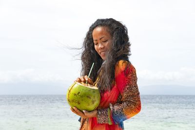 Young woman holding coconut water at beach against sky