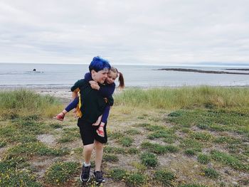 Happy brother piggybacking sister while standing on field by sea against cloudy sky