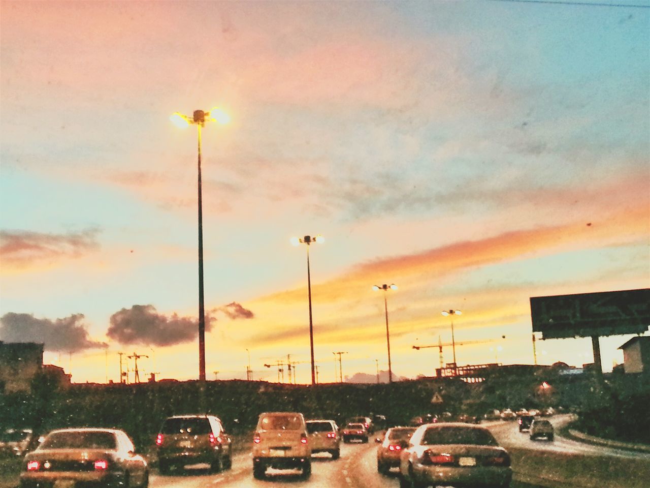 transportation, car, land vehicle, mode of transport, sunset, street light, sky, road, street, cloud - sky, traffic, parking, parking lot, stationary, orange color, city, on the move, parked, in a row, built structure