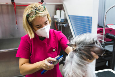 From above of anonymous female groomer in mask and uniform combing chinese crested dog with brush on grooming table during covid 19 pandemic